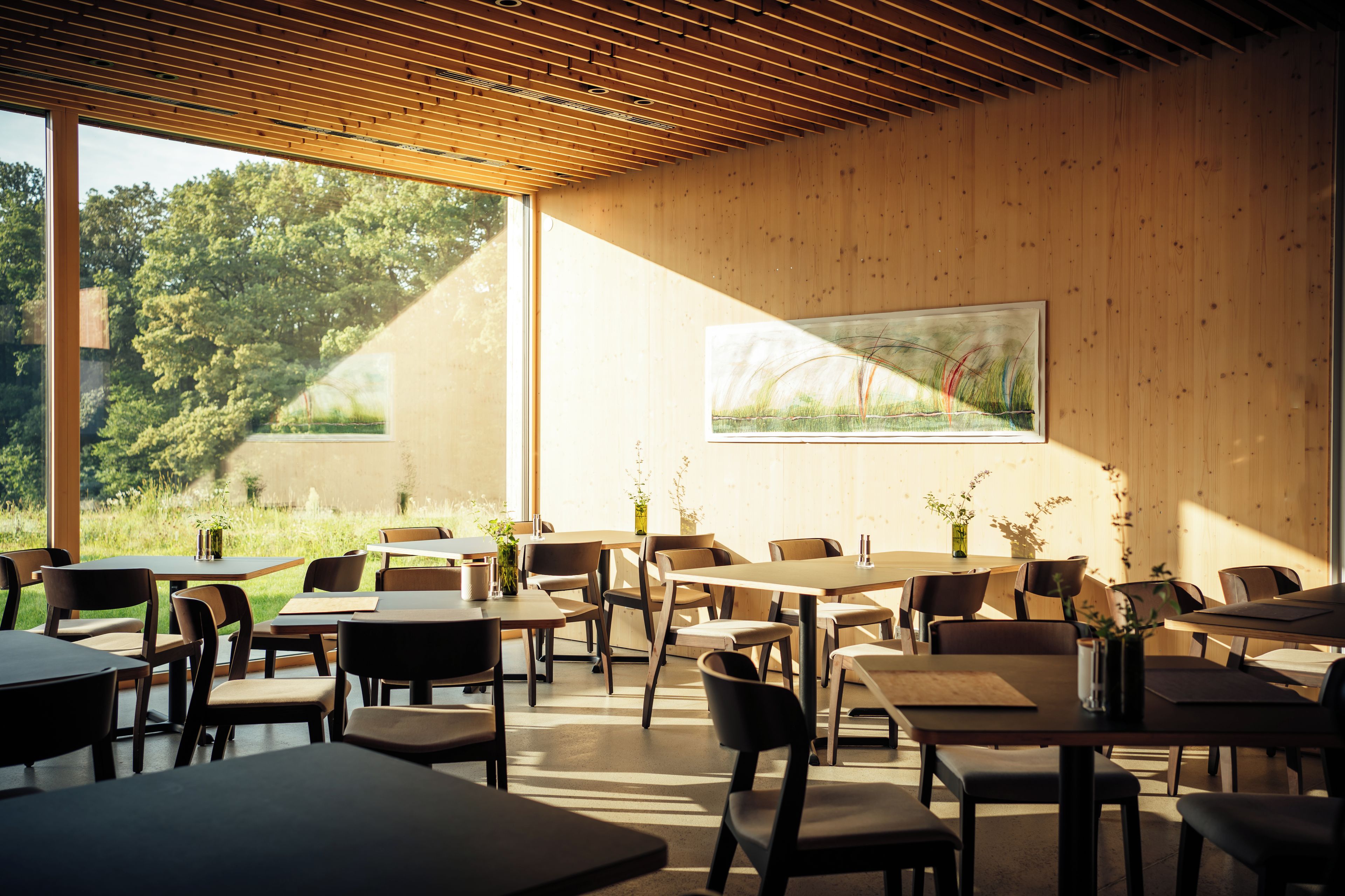 A modern interior with wooden ceiling beams, large windows, and a painting of grass. Tables and chairs are arranged for dining.