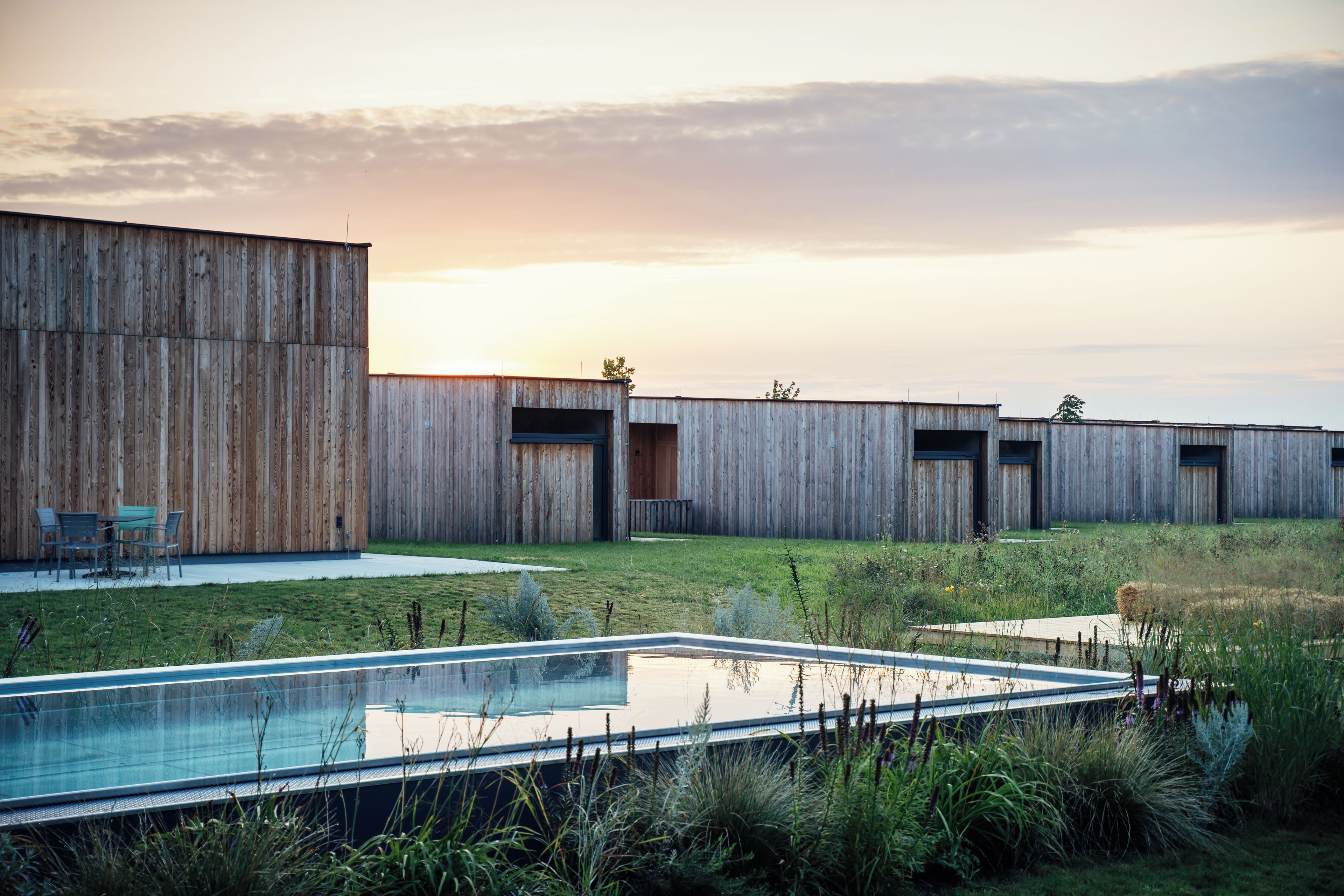 A modern building with a pool, surrounded by a grassy area and wooden structures.