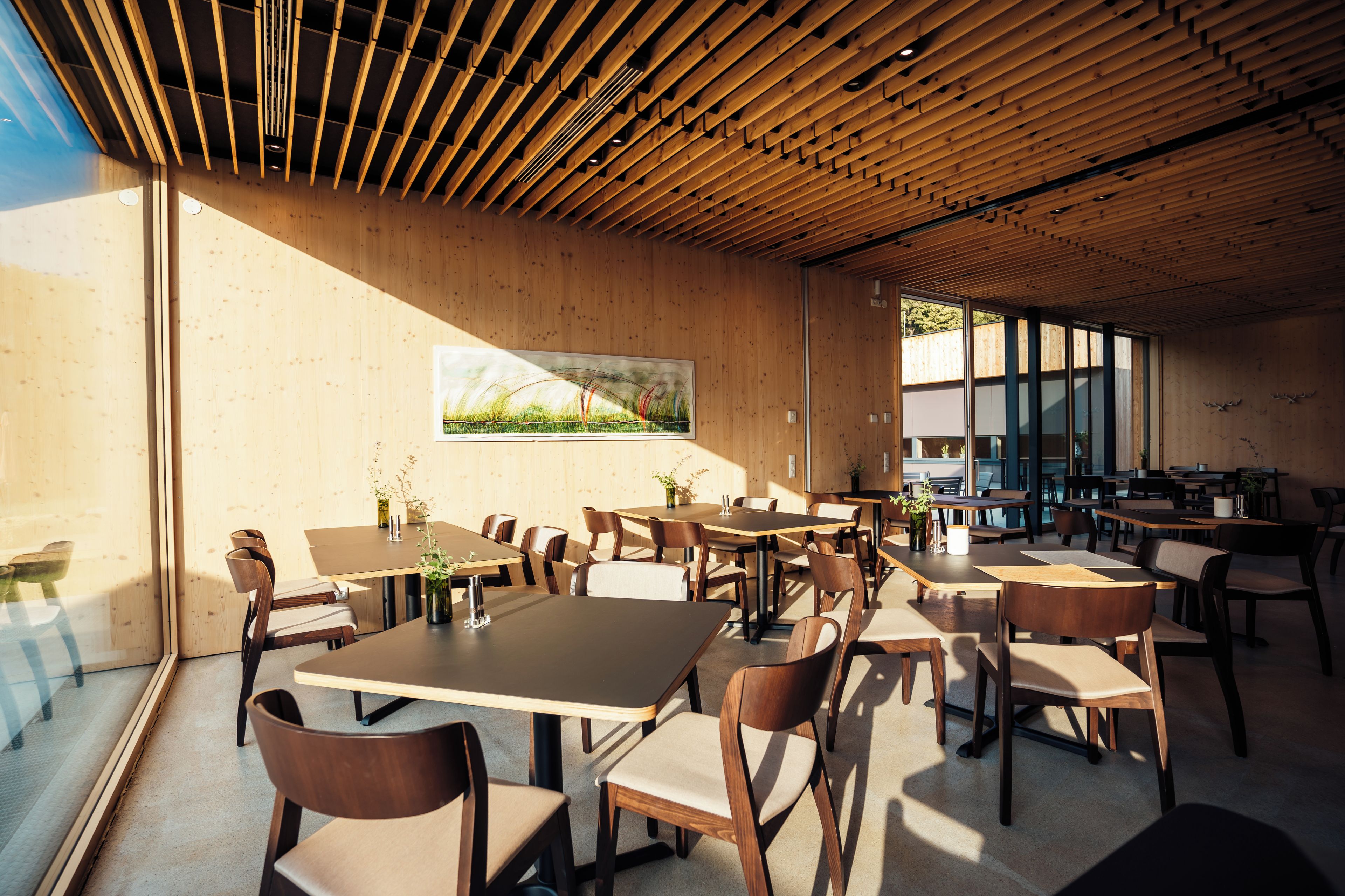A spacious dining area with multiple tables and chairs, wooden ceiling beams, and large windows allowing natural light.