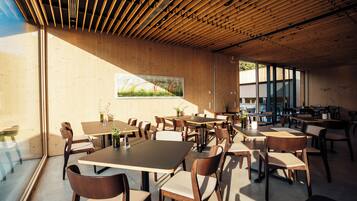 A spacious dining area with multiple tables and chairs, wooden ceiling beams, and large windows allowing natural light.
