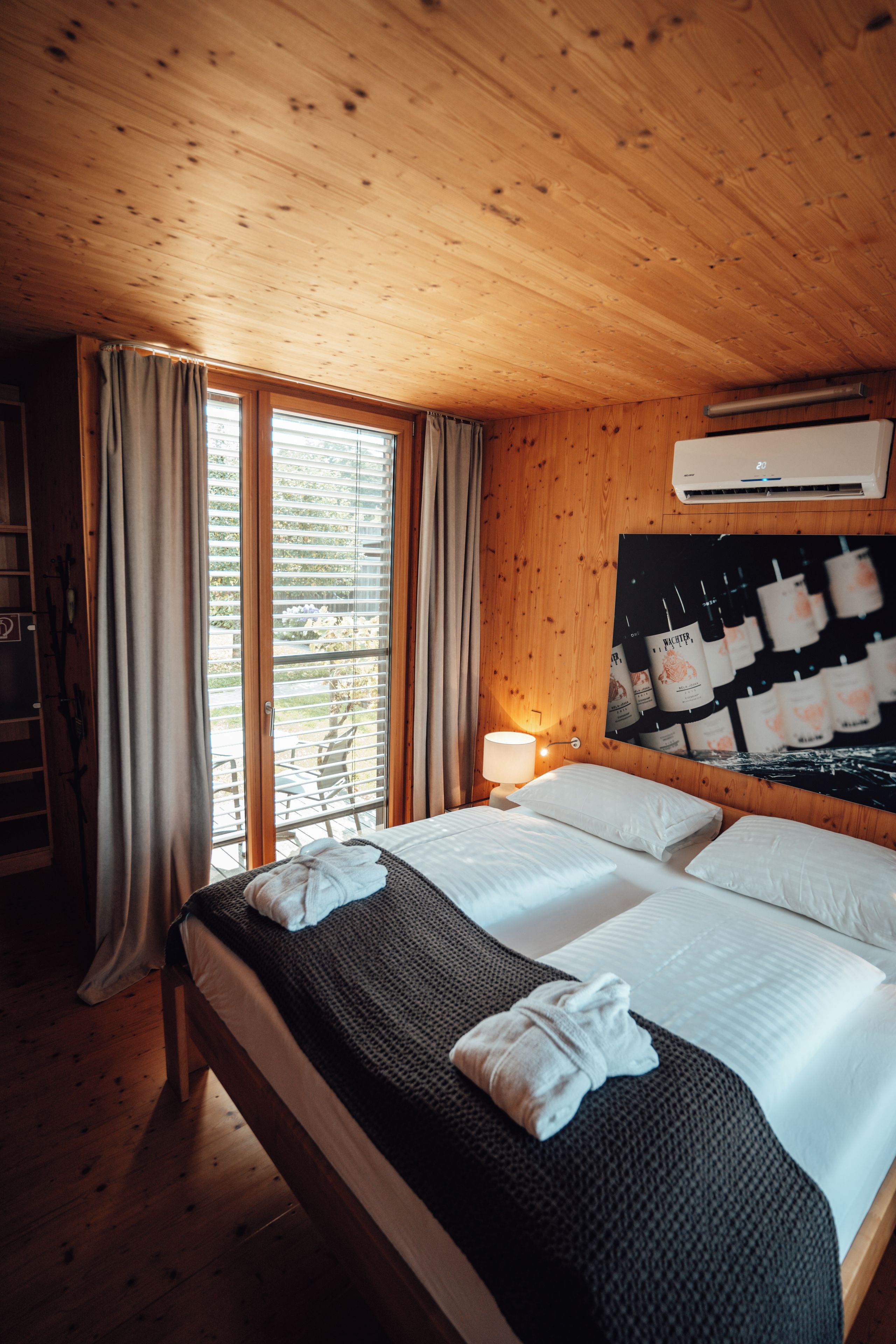 A wooden room with a bed, a window with curtains, a wall-mounted air conditioner, and a wall with framed pictures.