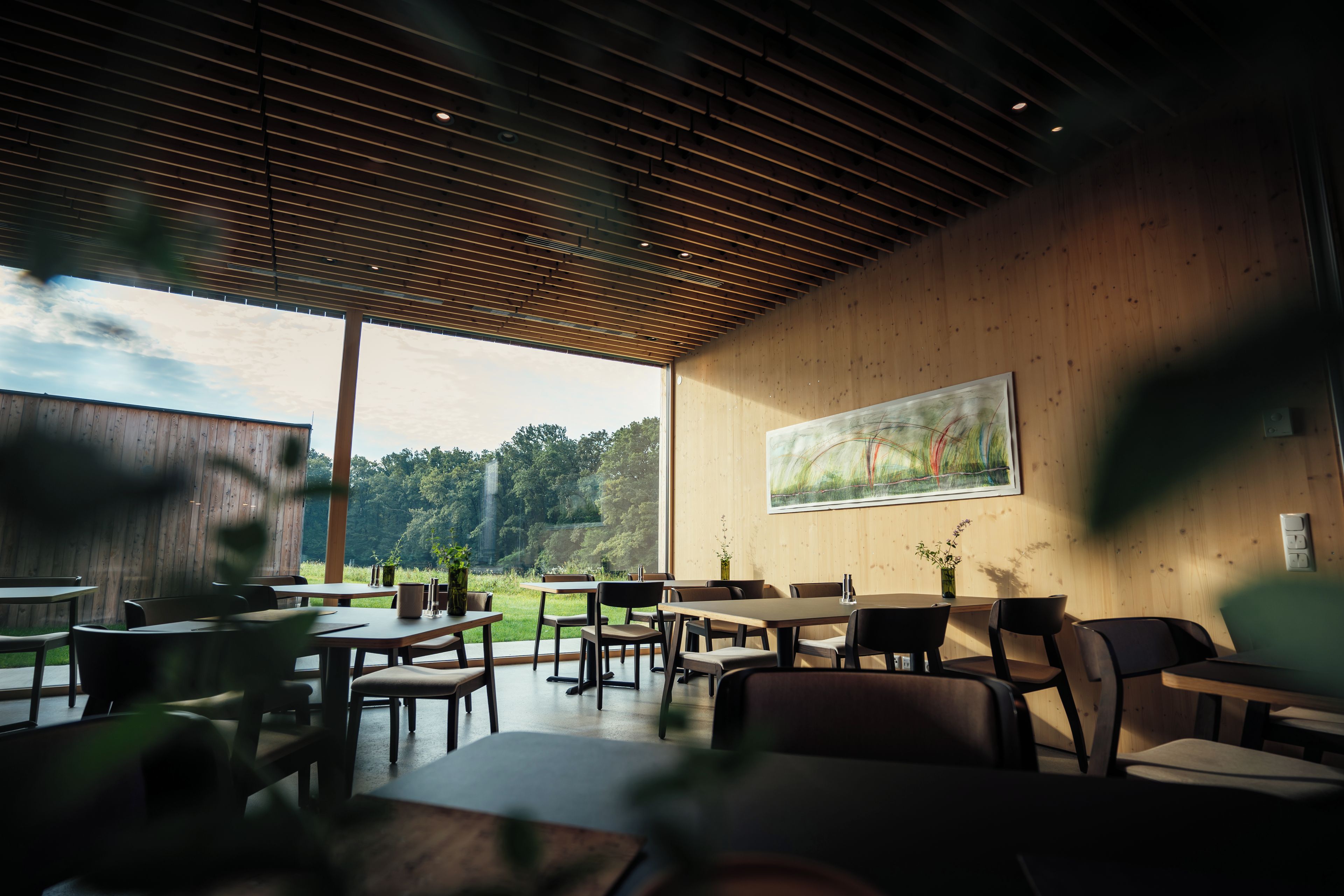 An indoor dining area with wooden walls and ceiling, tables, chairs, and a large window overlooking a garden.
