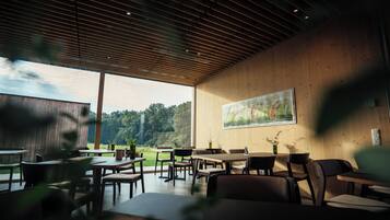 An indoor dining area with wooden walls and ceiling, tables, chairs, and a large window overlooking a garden.
