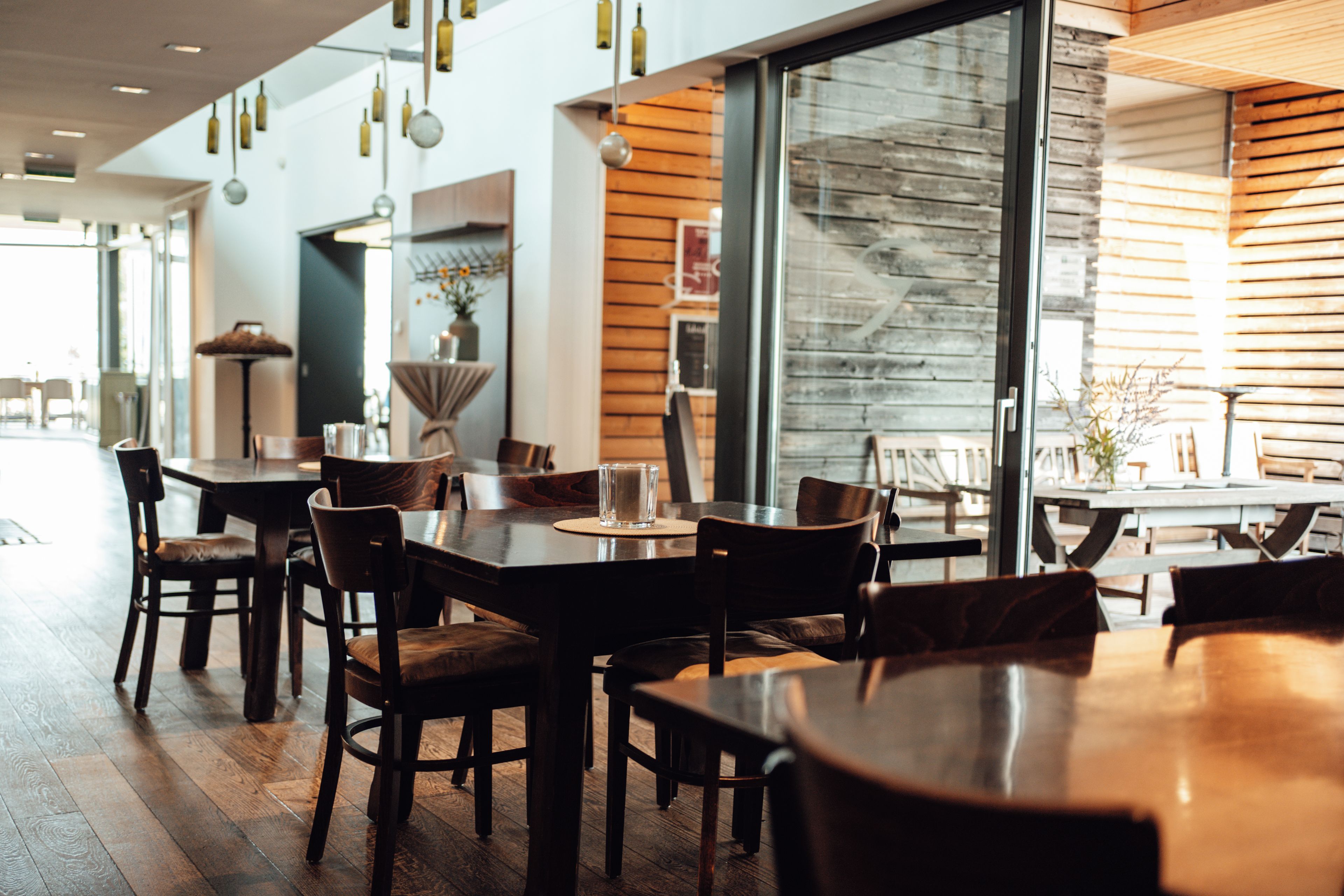 A modern restaurant interior with wooden tables and chairs, pendant lighting, and large windows.
