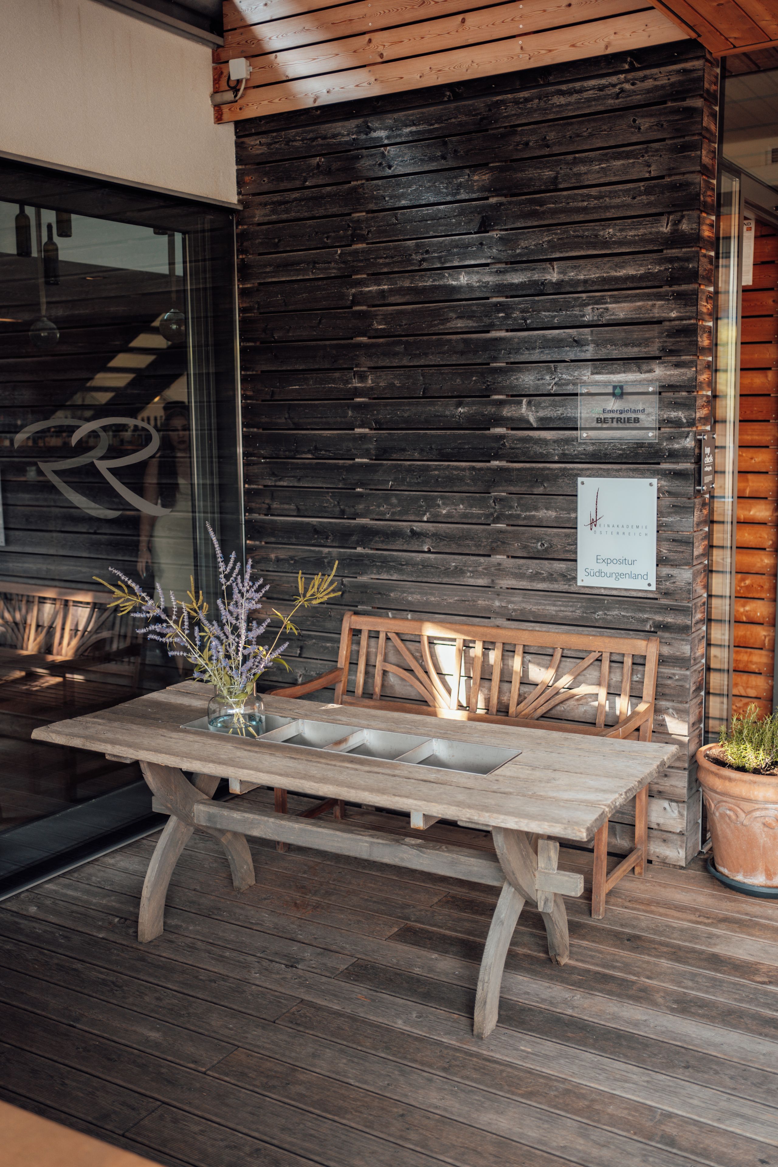 Una mesa de madera con un florero, un banco y una planta en maceta sobre una terraza de madera.