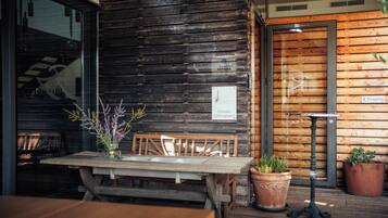 Pared exterior de madera con puerta de cristal, un banco de madera, una mesa con un jarrón de flores y dos plantas en macetas.