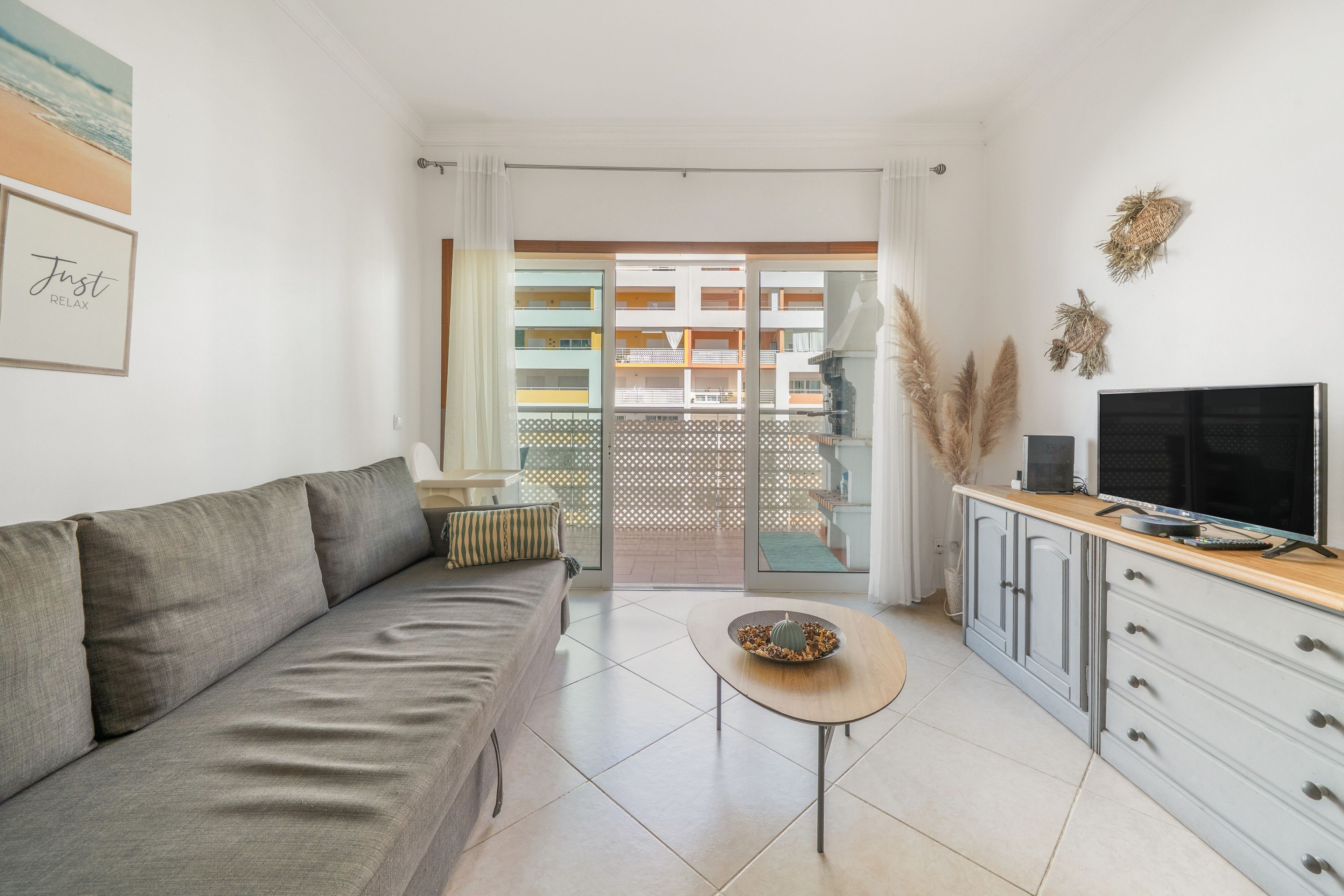 A modern living room with a grey sofa, a small round coffee table, a TV on a wooden cabinet, and a view of a balcony with a building in the background.