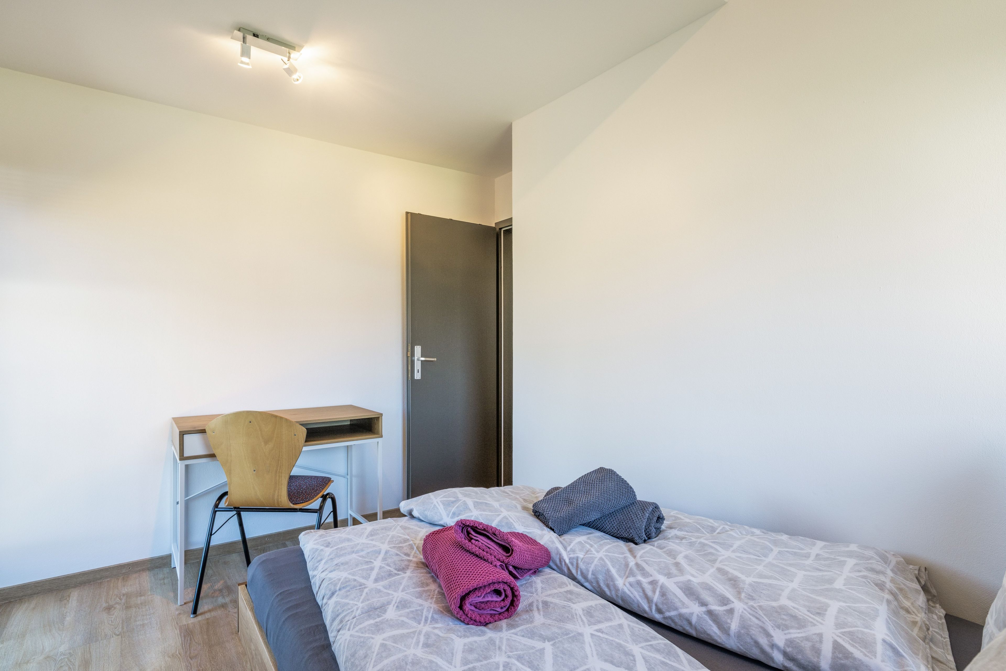 A small, modern room with a bed, a desk, and a chair.