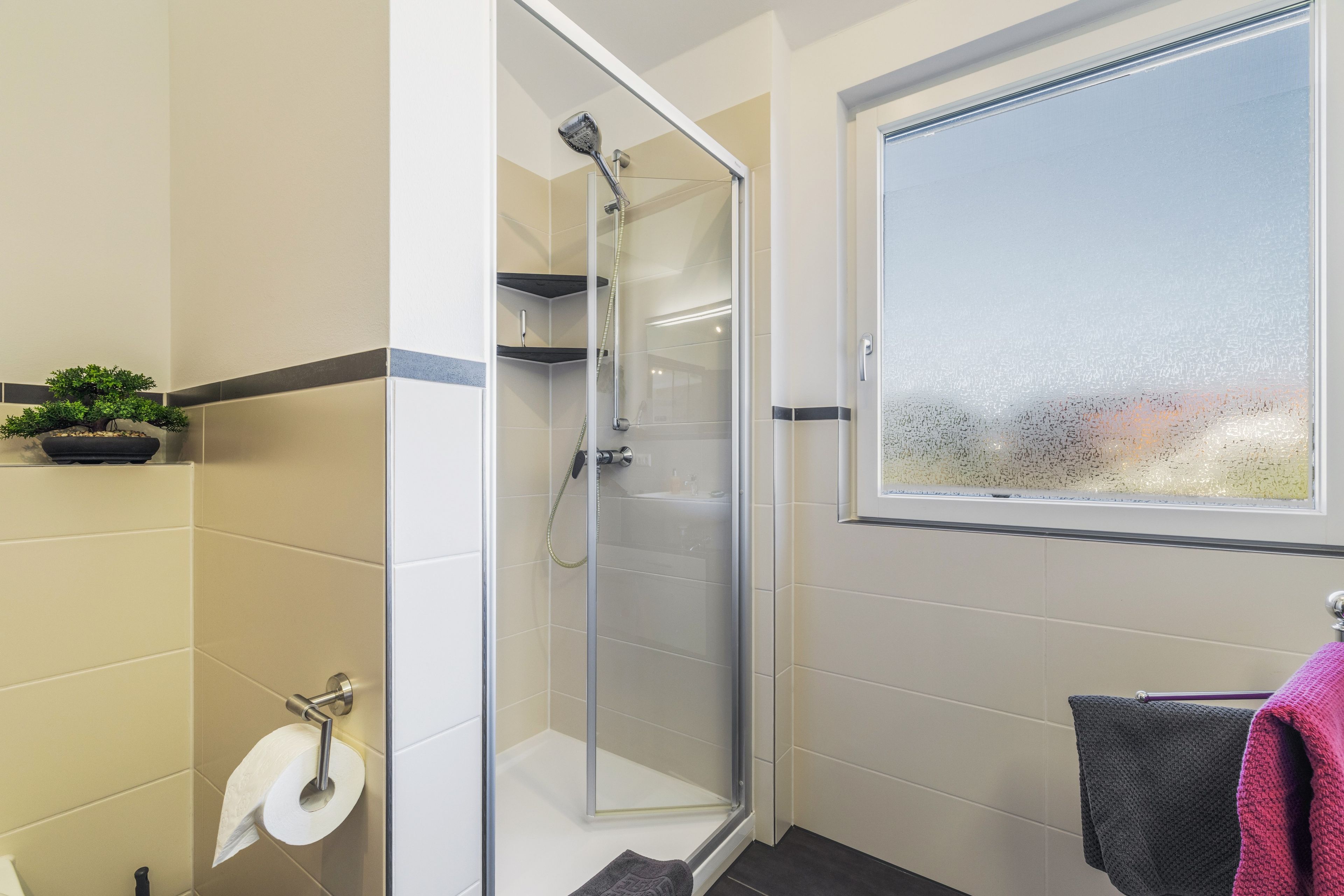 Ein modernes Badezimmer mit einer gläsernen Duschkabine, einem Toilettenpapierhalter, einem Fenster mit Blick und einem Handtuchhalter.