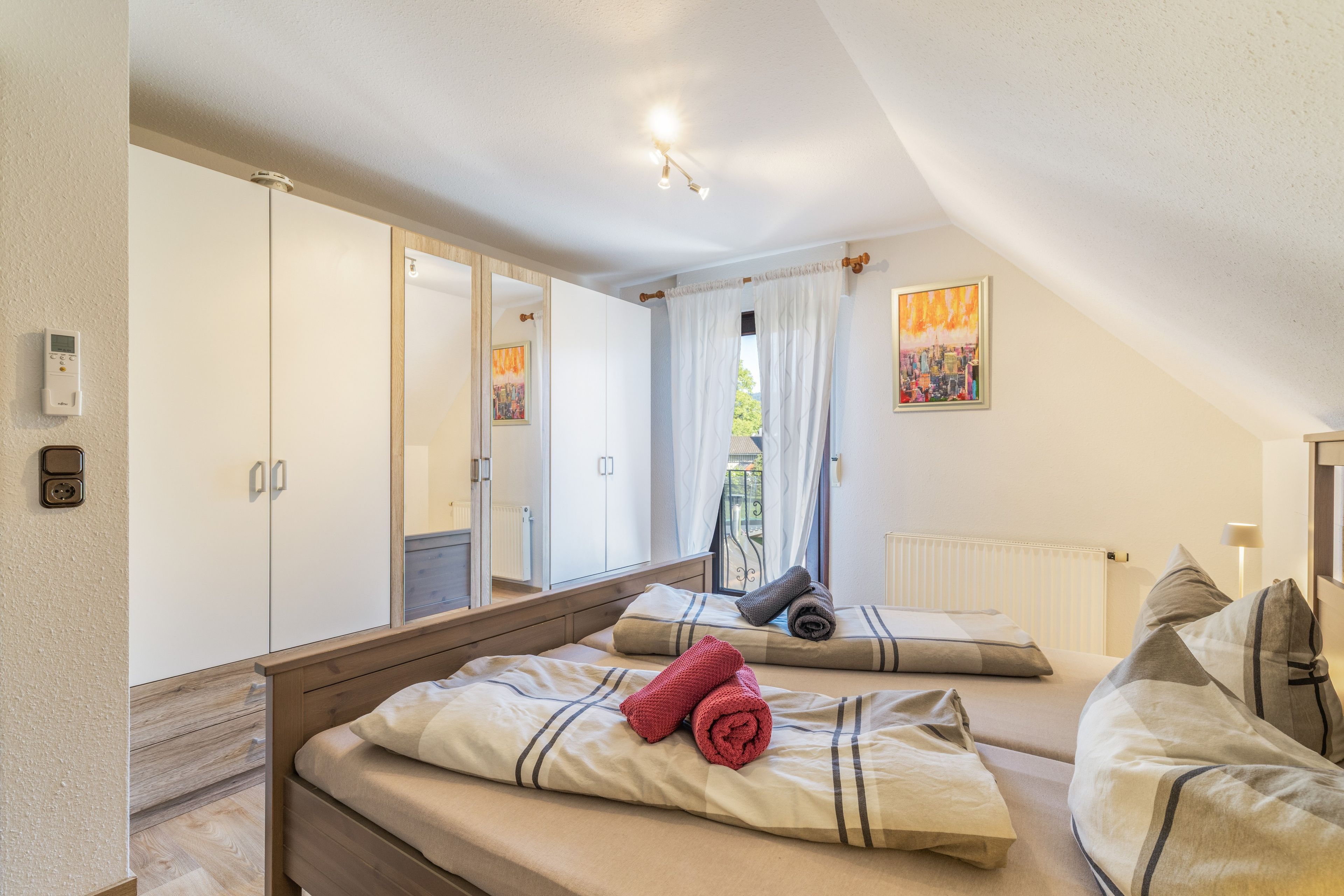 Ein Schlafzimmer mit Bett, Kleiderschrank, Spiegel, einem Gemälde und einem Fenster mit Vorhängen.