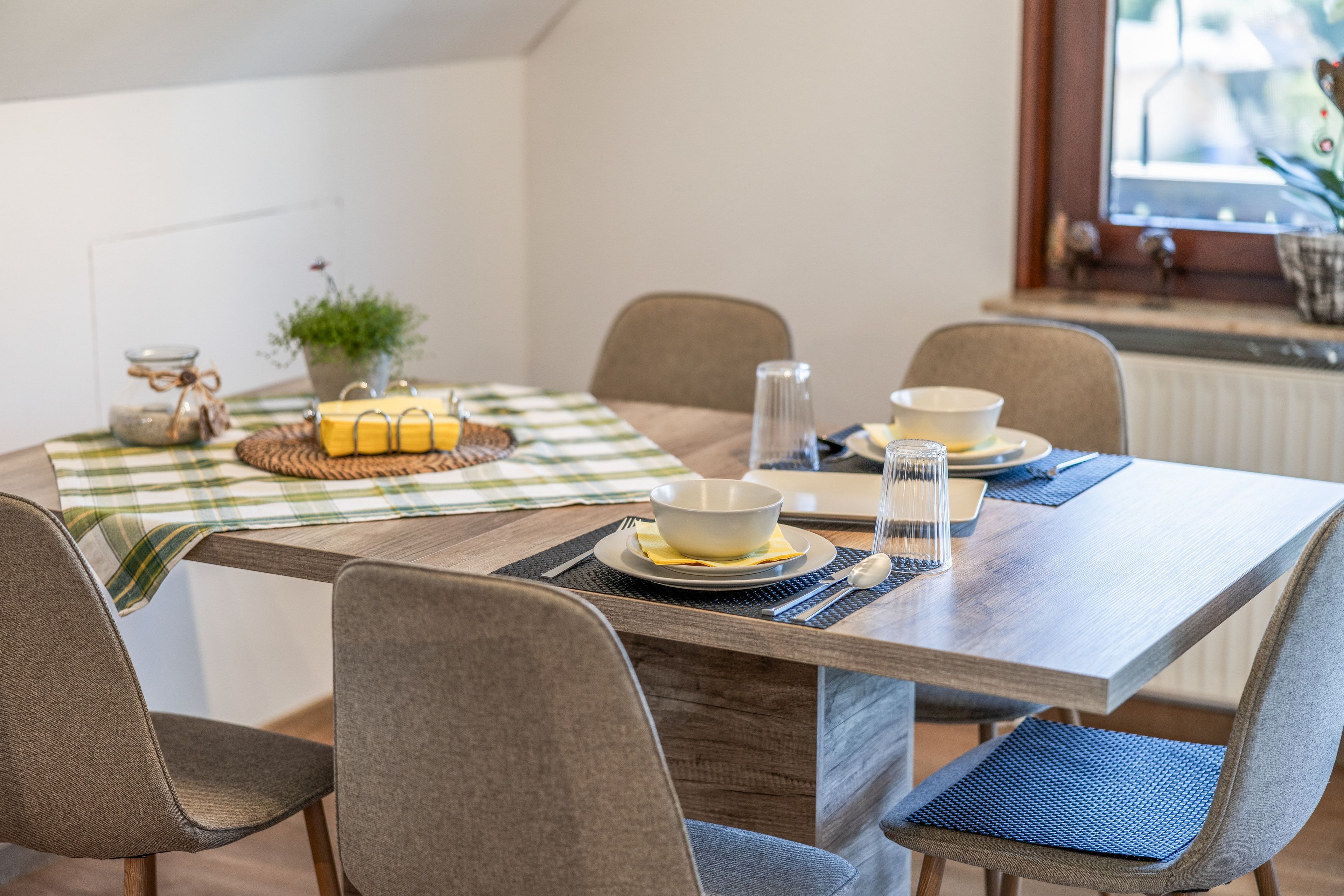 A dining area with a wooden table, four chairs, a checkered tablecloth, and a small plant.