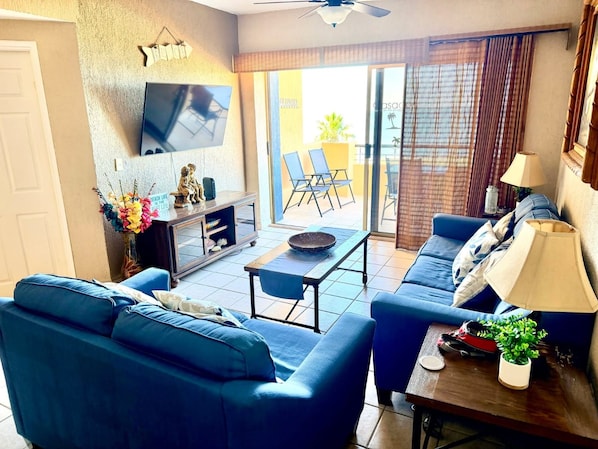 Un salon avec deux canapés bleus, une table basse, un téléviseur fixé au mur et une porte-fenêtre coulissante donnant sur un espace extérieur.