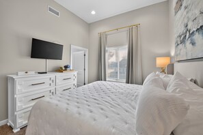 A bedroom with a bed, a dresser, a television, a window with curtains, and a painting on the wall. - Ivory Summit - Game Day, Concerts & Downtown! (Tuscaloosa)