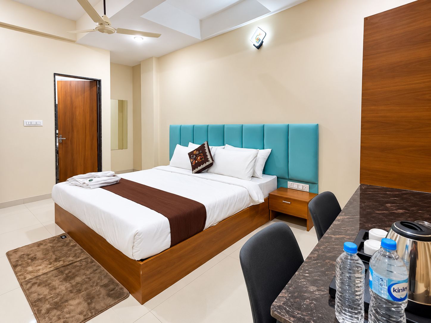 A hotel room with a bed, bedside table, chair, and a desk with a kettle and water bottles.