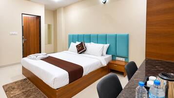 A hotel room with a bed, bedside table, chair, and a desk with a kettle and water bottles.