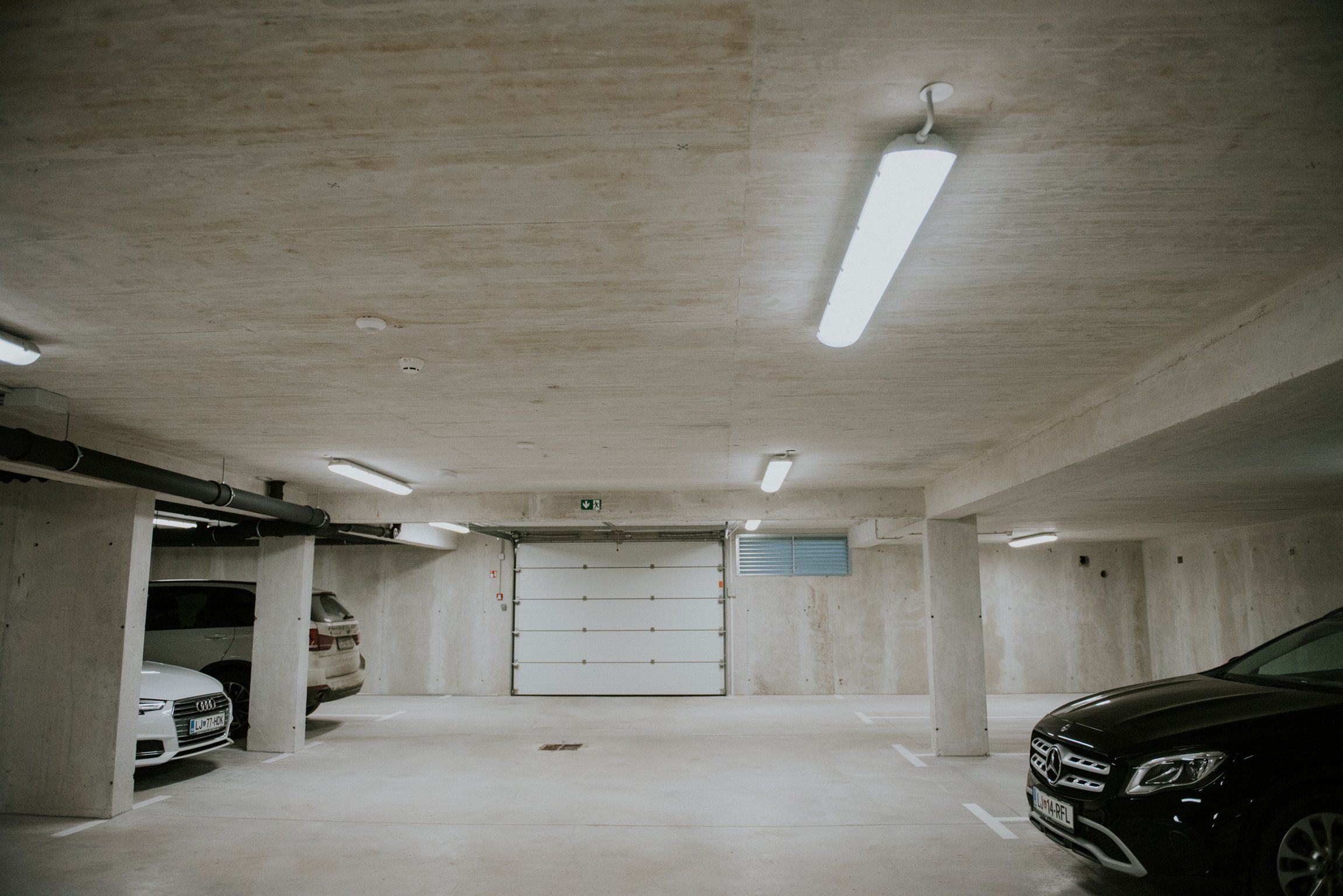 A parking garage with concrete pillars, fluorescent lighting, and a closed garage door.