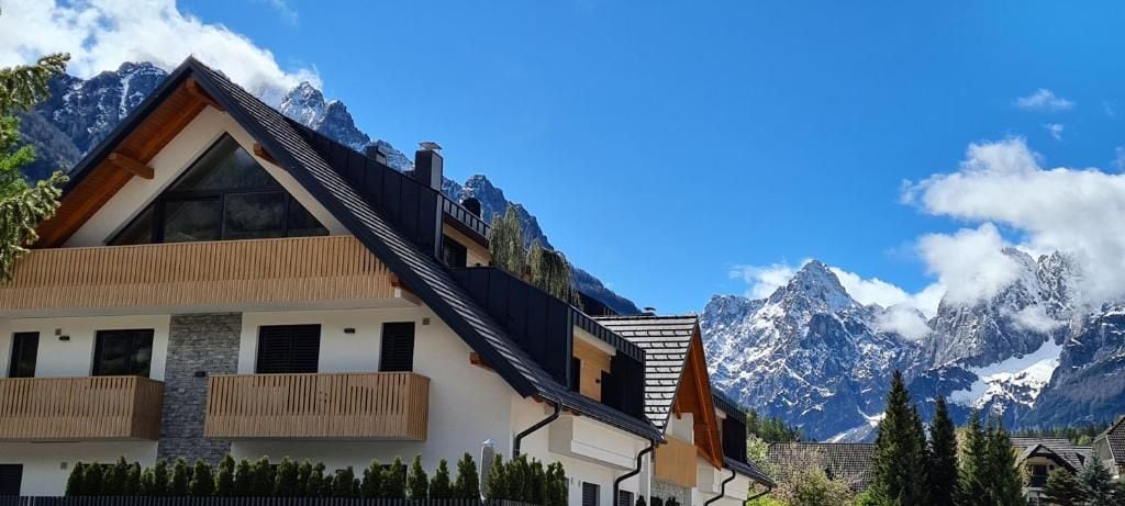 A modern two-story house with a sloped roof, large windows, and a balcony, set against a backdrop of snow-capped mountains.