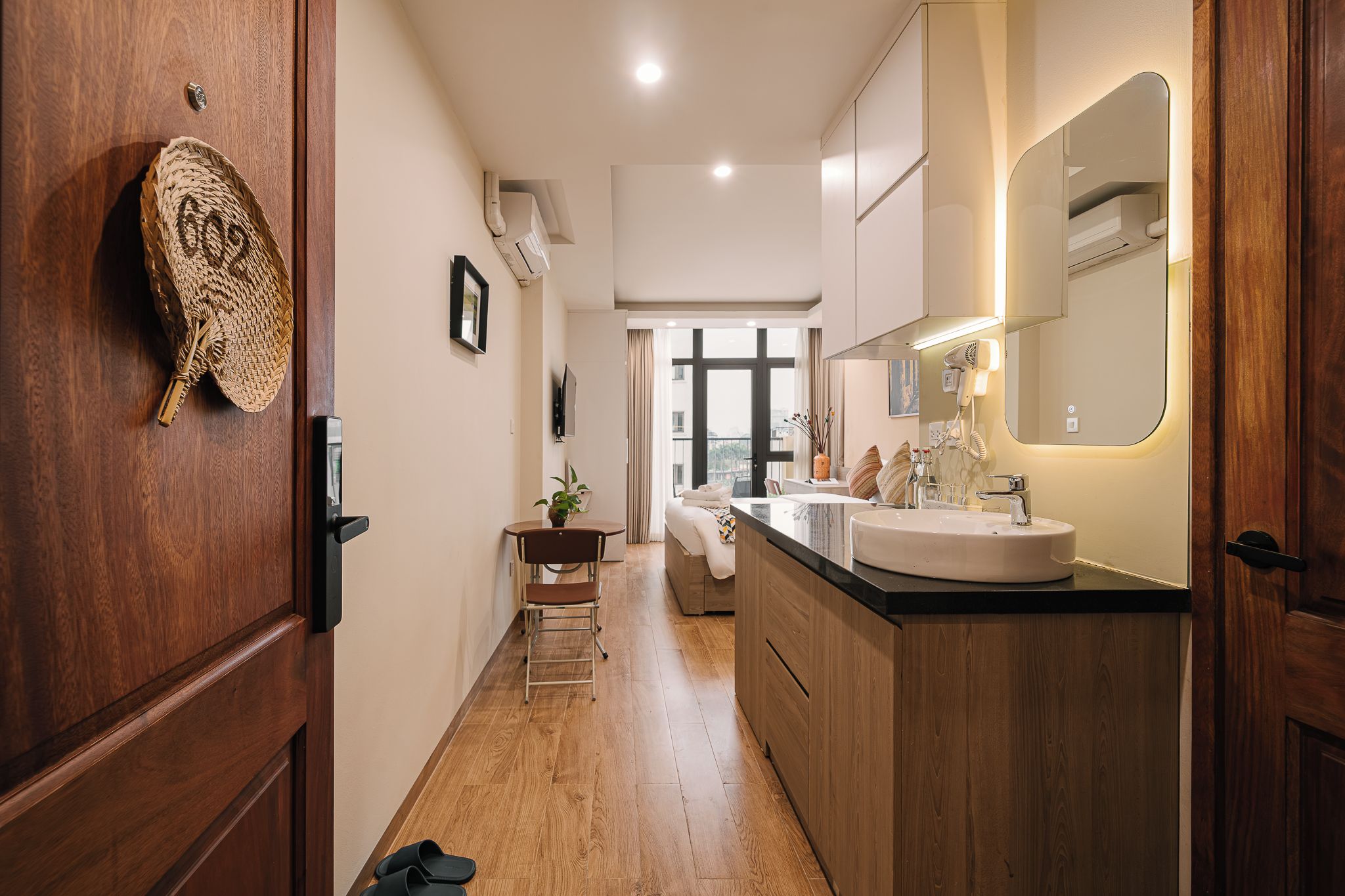 Ein Hotelkorridor mit Holzboden, einer Holztür mit gewebtem Teppich, einem kleinen Tisch mit Pflanze, einem Bett und einem Badezimmer mit Waschbecken und Spiegel.