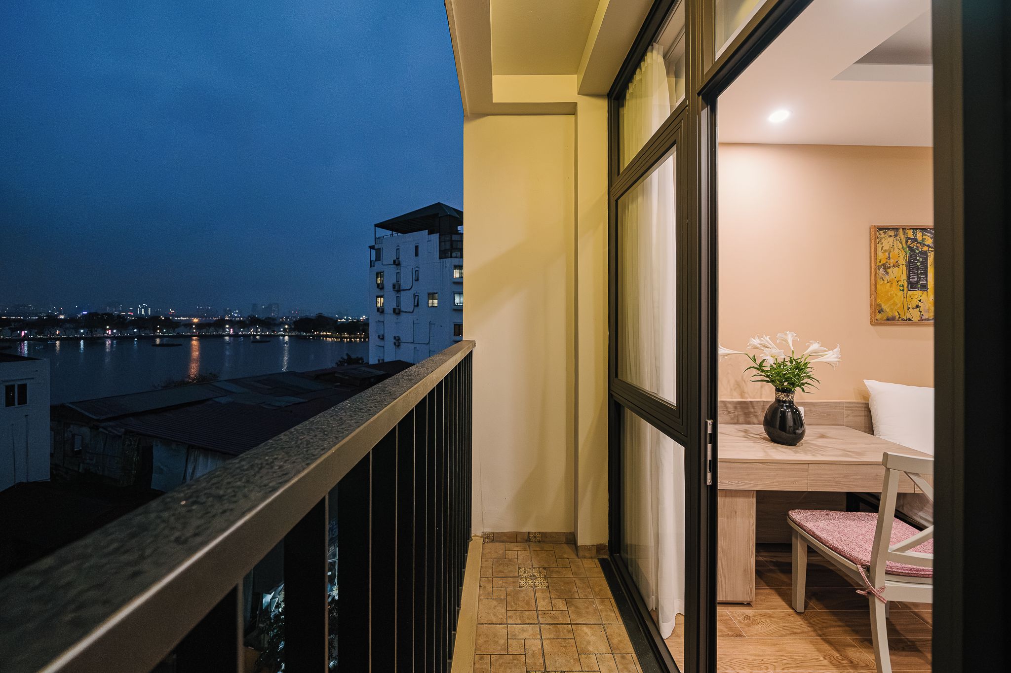 A balcony with a view of a cityscape at dusk, a dining area with a table and chairs, and a potted plant.