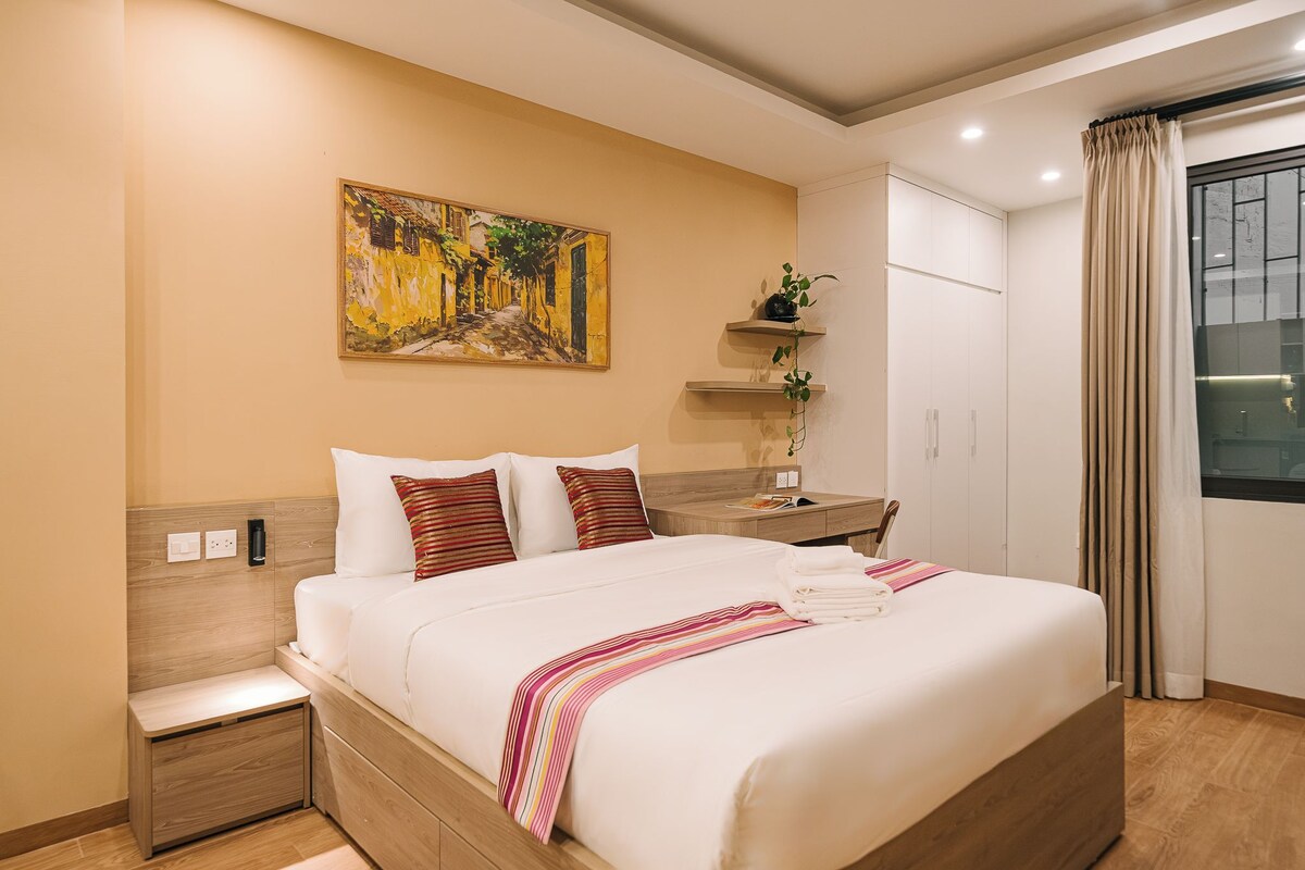 Une chambre d’hôtel avec un lit, une table de chevet, un tableau au mur, une plante et une fenêtre avec des rideaux.