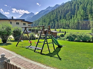 Ein Spielplatz mit Schaukeln und einer Rutsche, umgeben von einem gepflegten Rasen und einem Berg im Hintergrund.