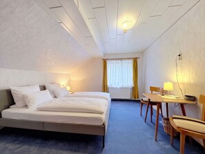 A hotel room with a bed, bedside table, lamp, chair, and a small table with a lamp.
