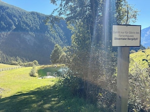 Ein Wegweiser mit Schrift in einer Fremdsprache, eine Rasenfläche, ein Gewässer und ein bewaldeter Berg im Hintergrund.
