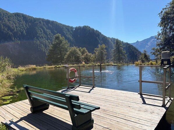 A wooden dock with a bench, a lifebuoy, and a water body surrounded by forested hills.