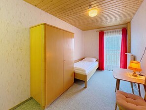 Ein kleines Zimmer mit einem Bett, einem Schrank, einem Tisch mit Lampe und einem fensterbepflanzten Fenster.