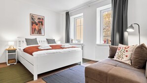 Une chambre à coucher moderne avec un lit blanc, un canapé marron, un tableau au mur et une fenêtre avec des rideaux.