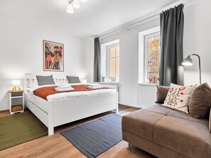Ein modernes Schlafzimmer mit einem weißen Bett, einem braunen Sofa, einem Bild an der Wand und einem Fenster mit Vorhängen.