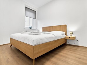 Ein modernes Schlafzimmer mit einem hölzernen Bett, weißer Bettwäsche, einem Nachttisch mit Lampe und einem Fenster mit Ausblick.