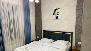 Cama doble con ropa de cama blanca y dos toallas, cabecera con adornos dorados, mesita de noche de madera y una obra de arte colgada en la pared.