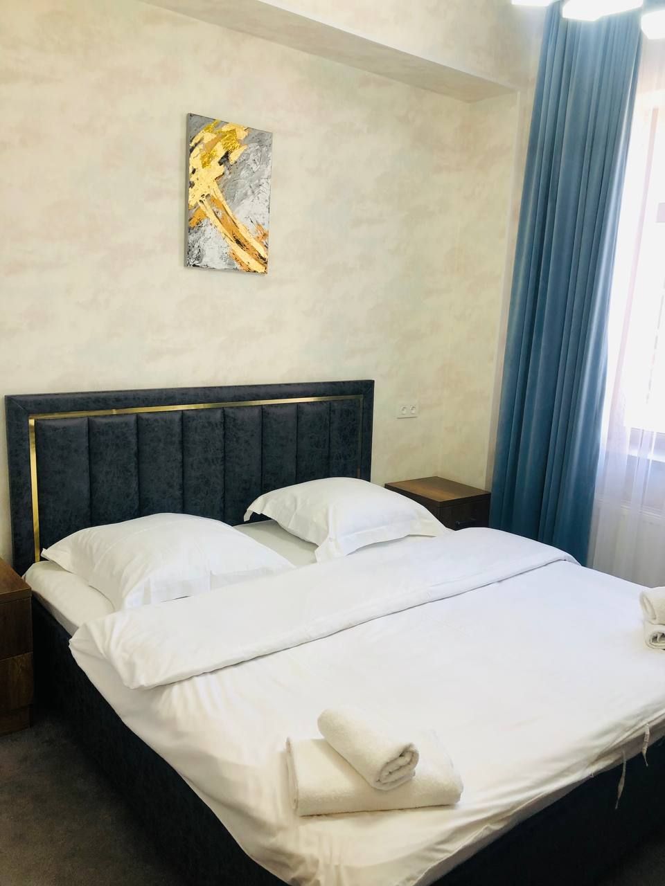 Una cama doble con ropa de cama blanca y una toalla doblada, una mesita de noche con lámpara, y un cuadro en la pared.
