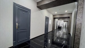 Un pasillo con dos puertas, una de ellas marcada con el número 212, que conduce a una habitación con suelo de baldosas negras y una zona de estar con sofá.