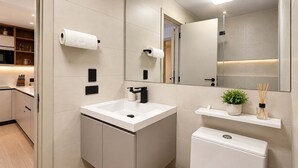 Un baño moderno con lavamanos blanco, grifo negro y un inodoro. Hay dos rollos de papel higiénico en la pared, un cubo de basura pequeño y un estante con una planta y difusor de aroma.