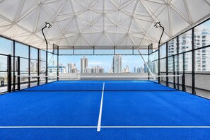 An indoor tennis court with a clear view of a city skyline through the netting. - 2BR at First del Sol Modern & Stylish (Panamá)