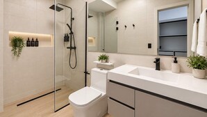 Un baño moderno con ducha de vidrio, inodoro blanco, lavabo con espejo y repisa con artículos de tocador.