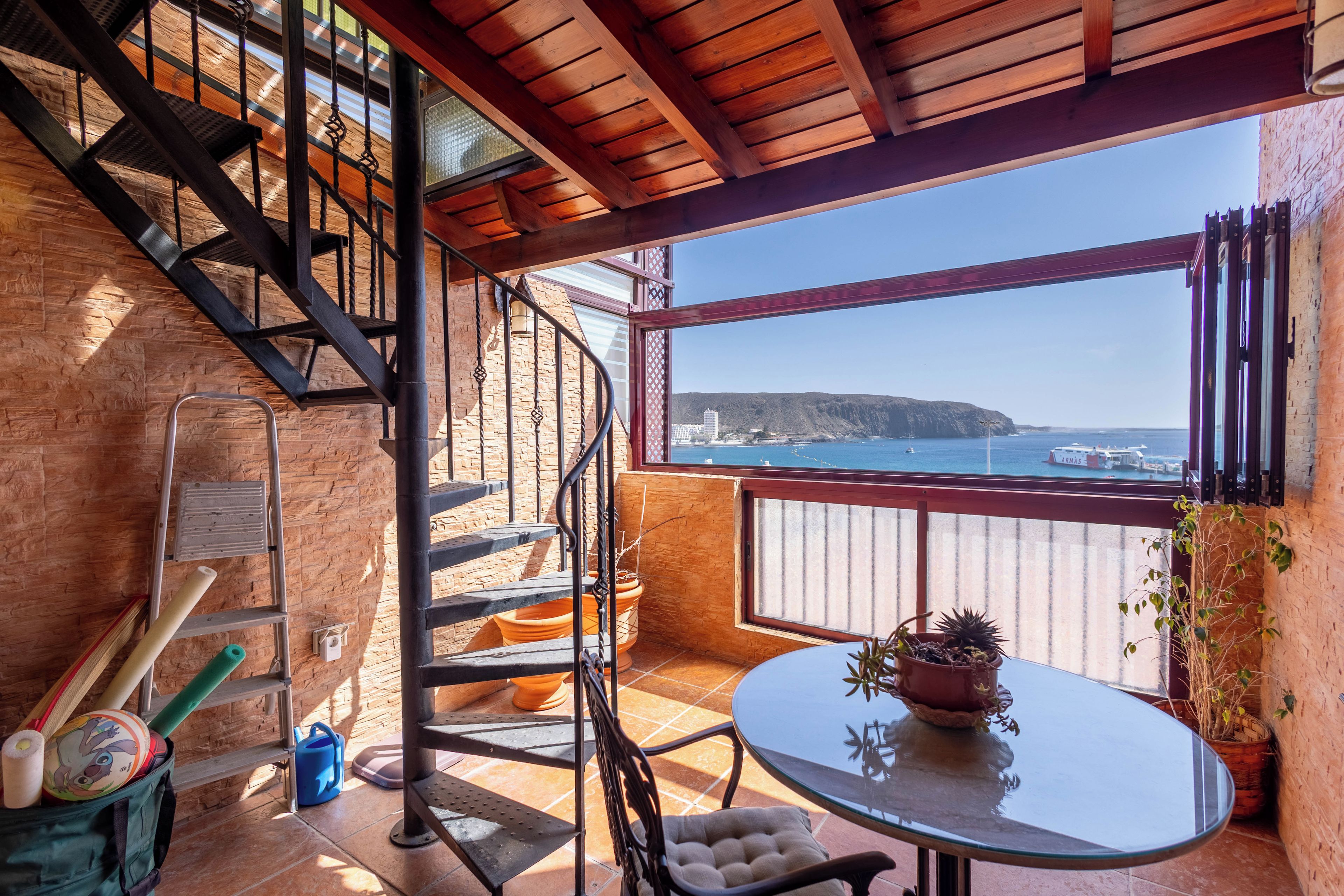 A balcony with a view of the sea, a round table, a chair, a potted plant, and a staircase.