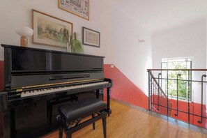 A black piano with a bench in a room with wooden flooring, a red wall, and a window with a view of greenery. - Contempora - Casa Osti - Designer Villa with Garden in the City Center (Milano)