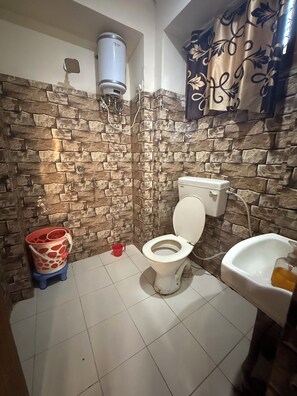A bathroom with a stone wall, a toilet, a red bucket, and a white sink. - Hotel Aditya Sheraton (Haridwar)
