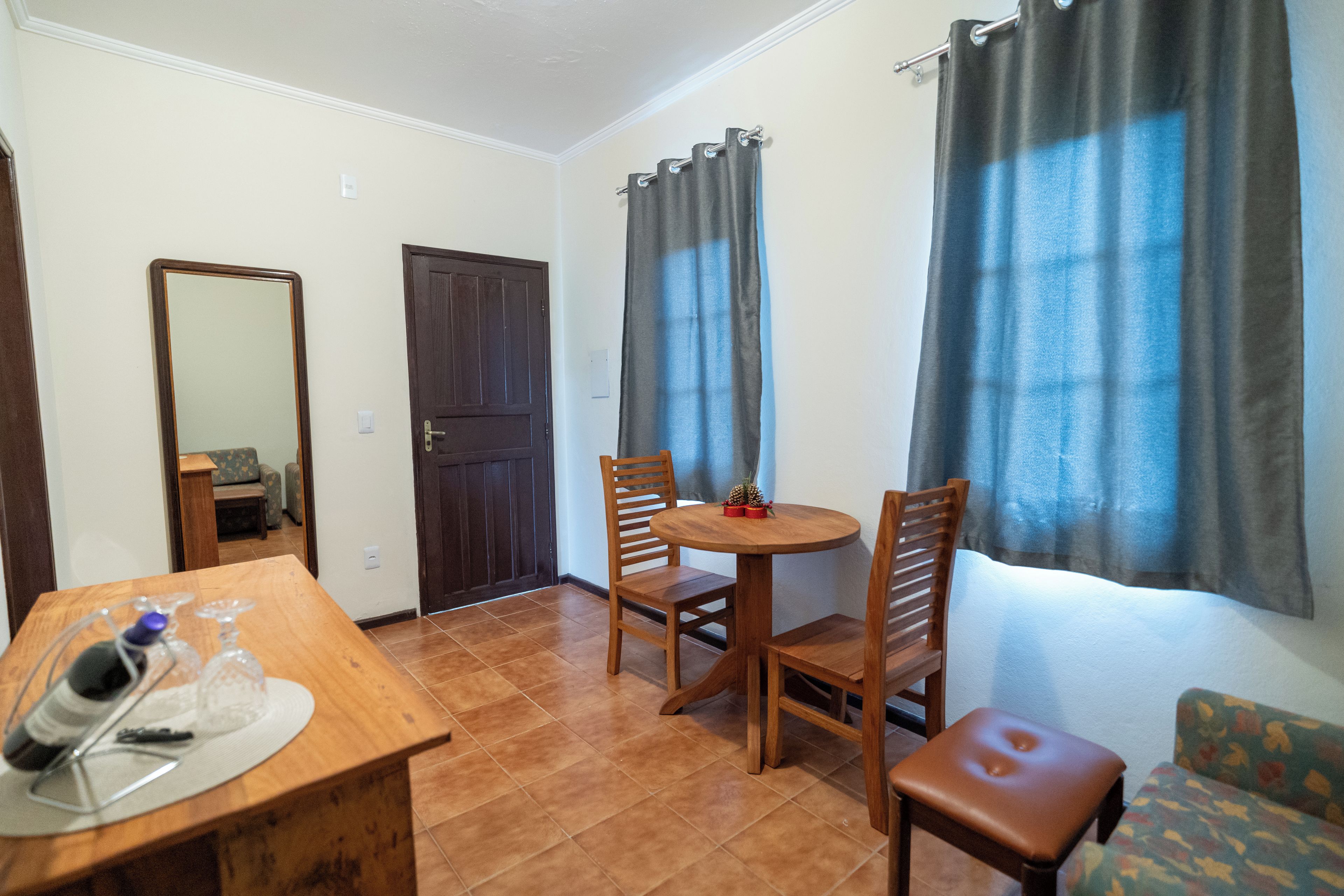 A room with a wooden dining table, two chairs, a small round table with a single chair, a sofa, a mirror, and a door.