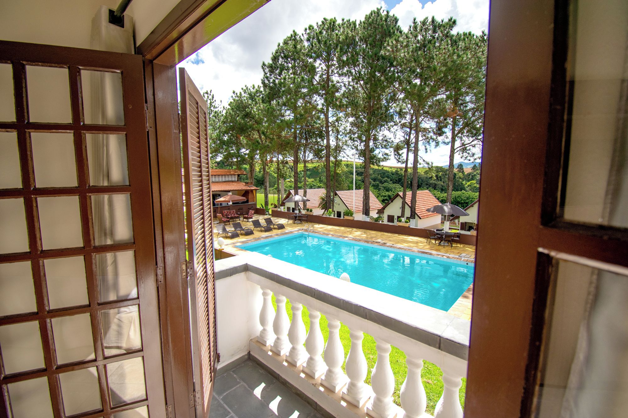 A view of a swimming pool with lounge chairs and umbrellas, surrounded by trees and a building with a red roof.