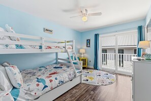 Quarto de criança com beliches, ventilador de teto, janela com cortinas e uma mesinha com abajur.