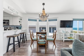 A modern kitchen with white cabinetry, a dining area with a glass table, and a large window overlooking the ocean. - 🌊La Mer | 3Br Oceanfront Condo with Shared Beach Walkover🧜‍♀️ (Carolina Beach)