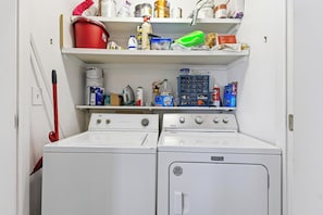 Uma lavanderia com duas máquinas de lavar e secar Maytag brancas, um esfregão vermelho e diversos produtos de limpeza nas prateleiras acima.