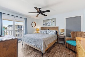 A hotel room with a bed, a ceiling fan, a window with a view of buildings, a chair, and a nightstand with a lamp. - 🌊La Mer | 3Br Oceanfront Condo with Shared Beach Walkover🧜‍♀️ (Carolina Beach)