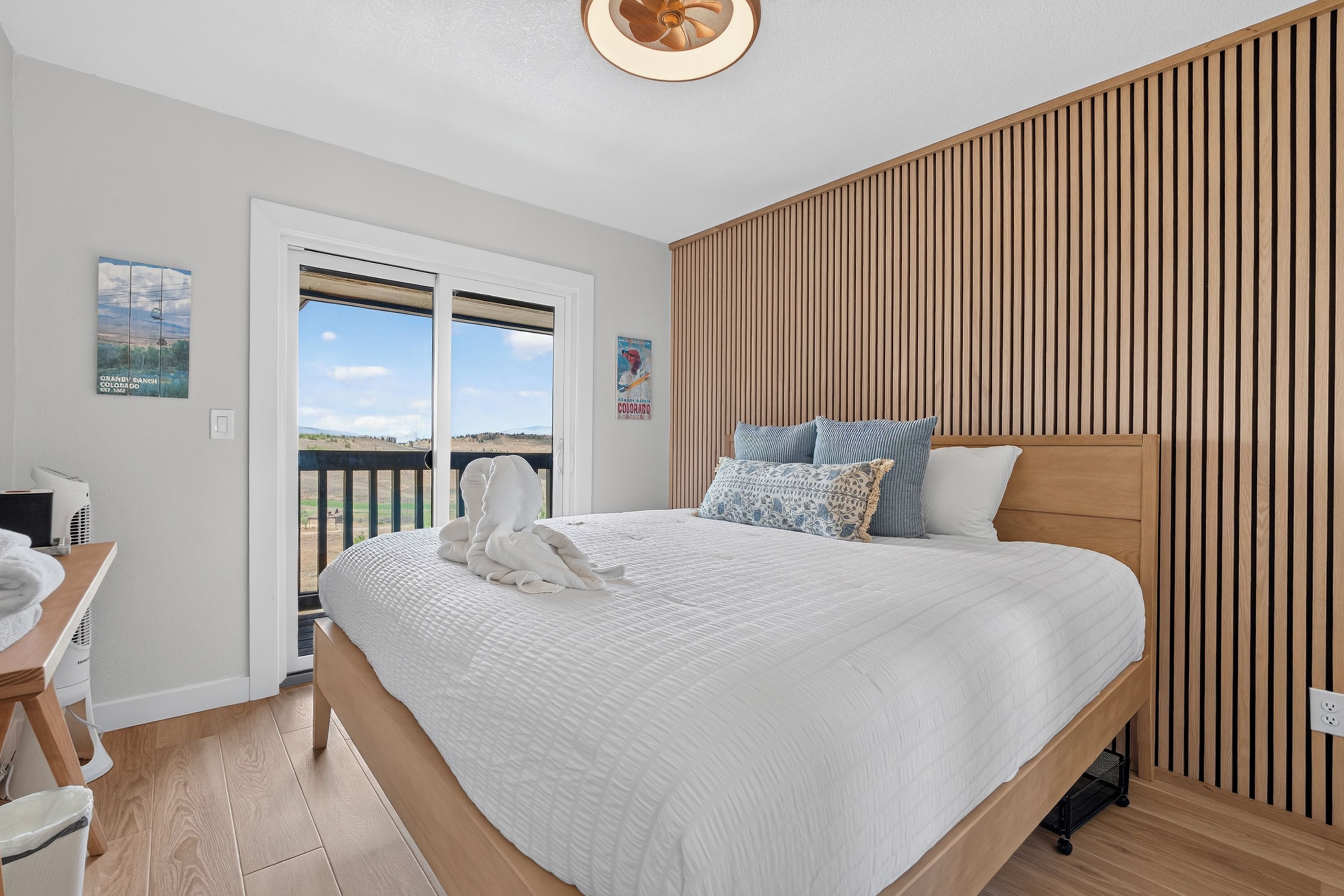 A bedroom with a large bed, a wooden slatted wall, a desk with a chair, and a view through a glass door.