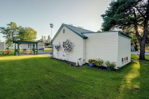 A small, single-story house with a green roof and white siding, surrounded by a well-maintained lawn and potted plants. - Walk to Lake Ontario! 'minnow Cottage' (Kent)