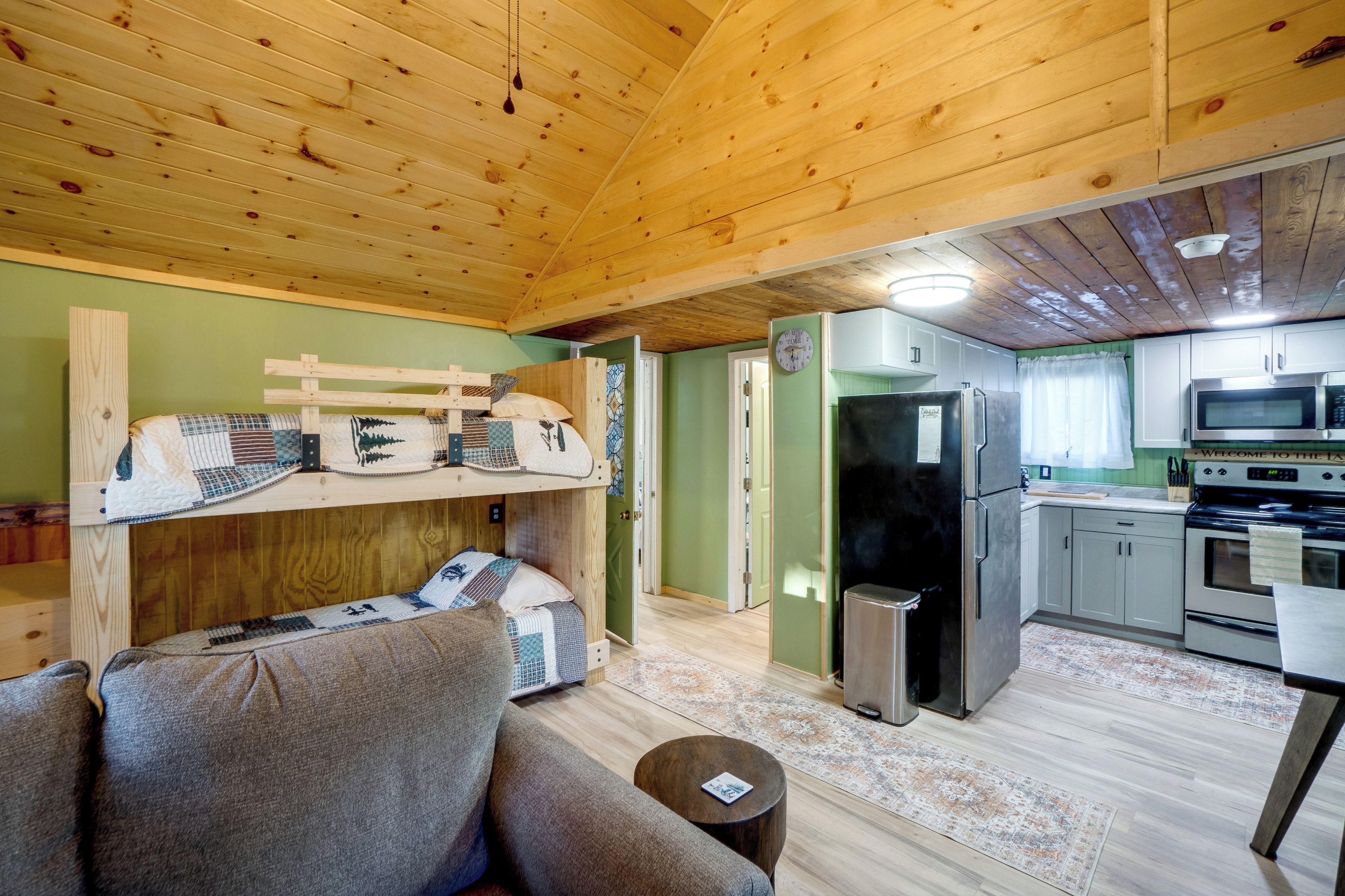 A cozy cabin interior with a bunk bed, a sofa, a kitchen with modern appliances, and a wooden ceiling.